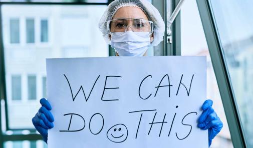 a healthcare work in ppe holds a sign that says we can do this