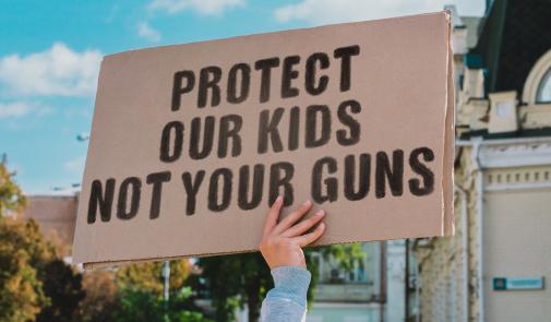 mano sosteniendo un cartel de cartón con letras negras que decían "protege a nuestros niños, no a tus armas"