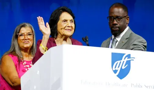 On the podium, Dolores Huerta waves to convention attendees, flanked by AFT officers Evelyn DeJesus and Fedrick Ingram.