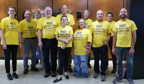 Twelve librarians in yellow shirts that read "No! Do not fire the librarianss! - Margaret Dunbar (WIU's 1st Librarian, 1902-1912)"