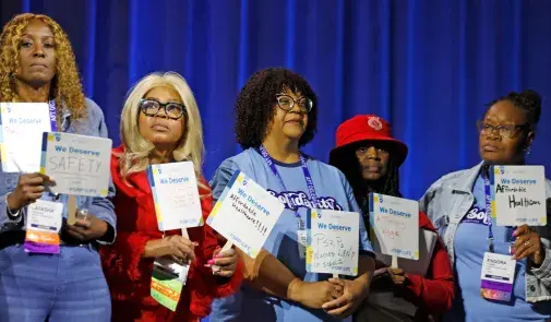Five PSRPs stand onstage with their fans bearing handwritten messages demanding safety on the job and healthcare.