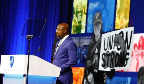 The Rev. Raphael Warnock, U.S. Senator from Georgia, stands at a podium in front of a images of AFT members and a sign that says "Union Strong"