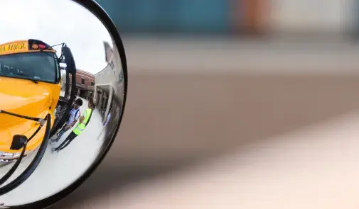 A reflection in a school bus mirror shows a bus driver helping a student board the bus.
