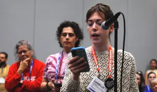 A woman holding a cell phone is speaking at a microphone while the audience listens.