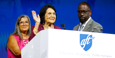 En el podio, Dolores Huerta saluda a los asistentes a la convención, flanqueada por los dirigentes de la AFT, Evelyn DeJesus y Fedrick Ingram.