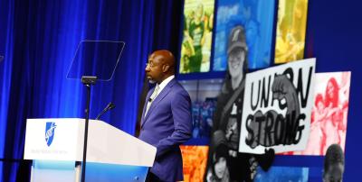El reverendo Raphael Warnock, senador estadounidense por Georgia, se encuentra en un podio frente a imágenes de miembros de la AFT y un letrero que dice "Unión Fuerte".