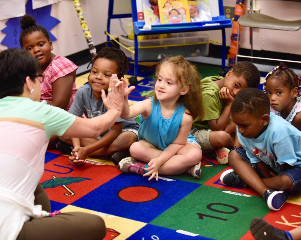 Photo of children sitting in a classroom and one giving a high five to a teacher