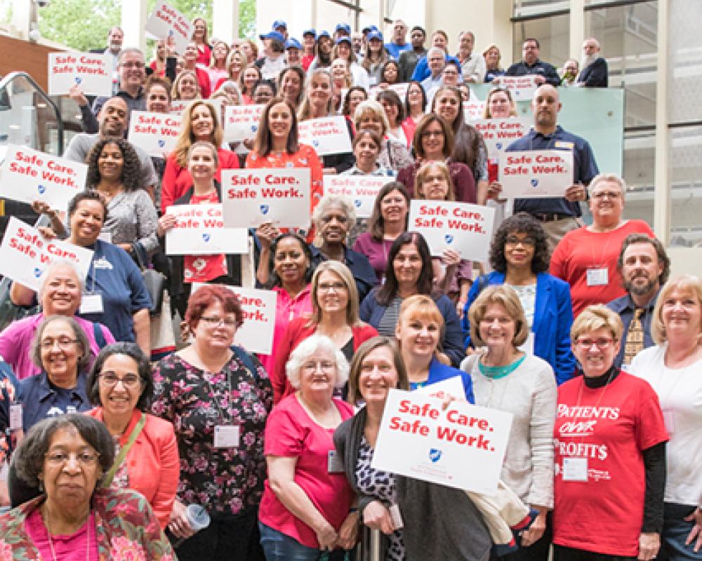  a group of people, mostly women, are standing, some with signs that say "safe care, safe work"