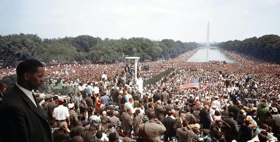 image of the 1963 March on Washington, MLK Jr in the bottom left corner in front of a massive crowd, the Washington Monument in the far distance across the National Mall