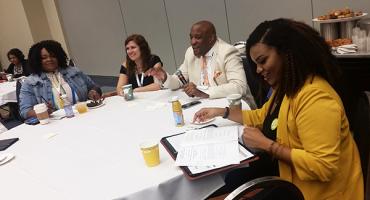 six people, five of whom are african american, discuss something at a table.