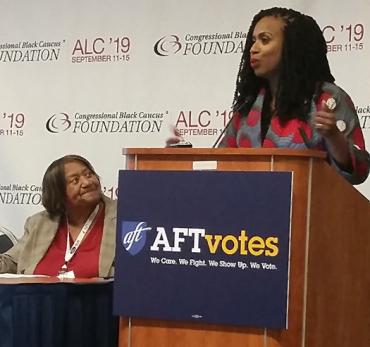 A black woman, Ayanna Pressley, speaks at a podium while an older black woman, Lorretta Johnsom, looks at her.