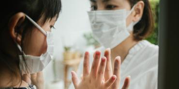 woman in mask holds up hand to a child in mask who is on the other side of a window