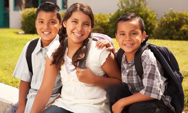 three latinx children smile while wearing backpacks