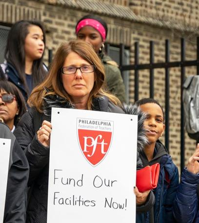 a woman holds a sign at the pft rally demanding that their facilities be funded