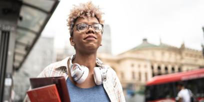 black woman with glasses holds books, looks toward the sky, has headphones resting around her neck