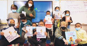 Randi Weingarten sits on the floor surrounded by school children and their teacher, holding picture books