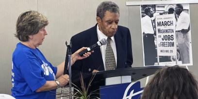 Phot of civil rights organizer Norm Hill speaking at the AFT breakfast. AFT President Randi Weingarten stands on the side.