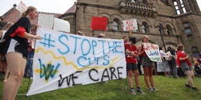 Estudiantes, profesores y empleados de la Universidad de West Virginia con carteles. El letrero en primer plano dice "#Stop the Cuts. We Care".