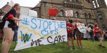 West Virginia University students, faculty, and employees holding signs. The sign in the foreground reads "#Stop the Cuts. We Care."