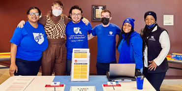 six adults behind a table covered in handouts. the adults are smiling with their arms around each other as they pose for the picture