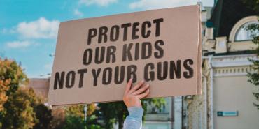 hand holding a cardboard sign with black letters reading "protect our kids not your guns"