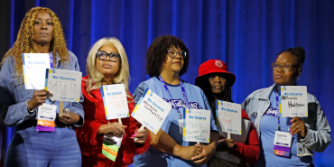 Five PSRPs stand onstage with their fans bearing handwritten messages demanding safety on the job and healthcare.