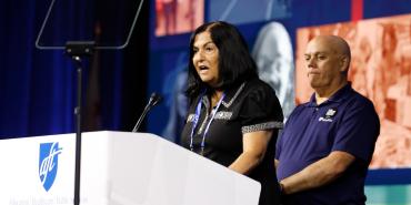 Anne Goldman stands at the podium with John Brady in the background.