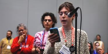 A woman holding a cell phone is speaking at a microphone while the audience listens.
