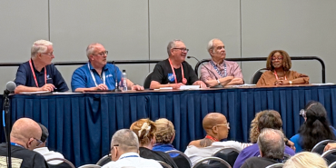 A group of people sit on a panel before a crowd.