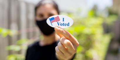 woman in coronavirus mask holds a sticker that says quote i voted unquote