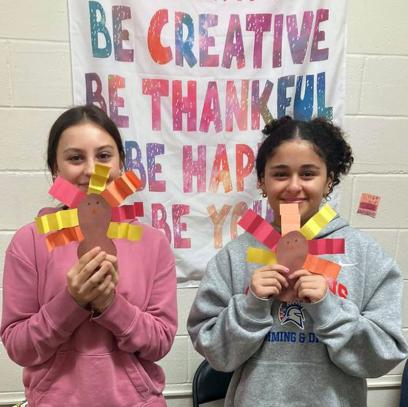 Foto de dos estudiantes haciendo manualidades de acción de gracias. El letrero detrás de ellos dice "Sea creativo. Sea agradecido. Sea feliz. Sea usted mismo".