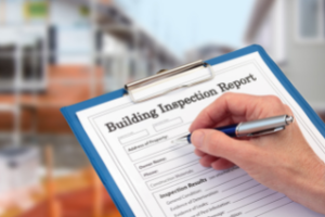 Foto de una persona sosteniendo un portapapeles y escribiendo en un formulario que dice "Informe de inspección del edificio"